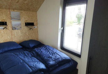 Kleines Schlafzimmer mit blauer Bettwäsche und großem Fenster im Solo Retreat Tiny House, Reestervallei, Niederlande.