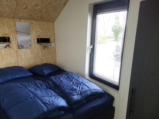 Pequeño dormitorio con ropa de cama azul y gran ventana en Solo Retreat tiny house en Reestervallei, Países Bajos.