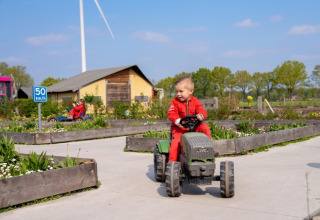 Een kind in een rode overall rijdt met een speelgoedtractor op een vakantieboerderij met windmolen en planten.