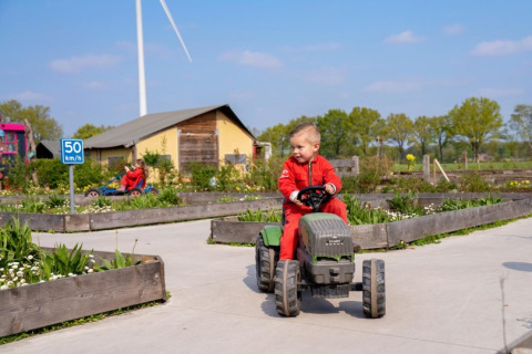 Een kind in een rode overall rijdt met een speelgoedtractor op een vakantieboerderij met windmolen en planten.