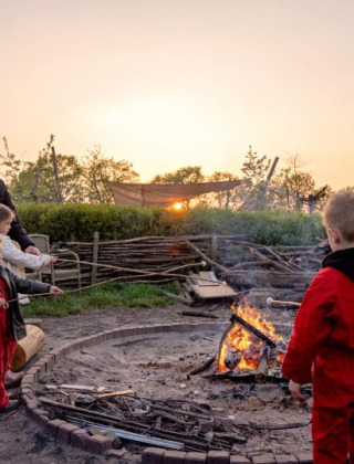 Niños y adultos asan malvaviscos al atardecer junto a una fogata en FarmCamps BoeBaDoe, Limburg, Países Bajos.