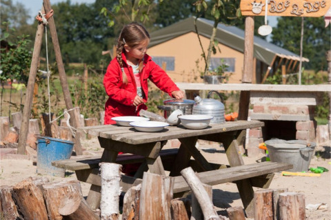 Mädchen im roten Anzug spielt draußen an einem Holztisch im FarmCamps BoeBaDoe in Limburg, Niederlande.