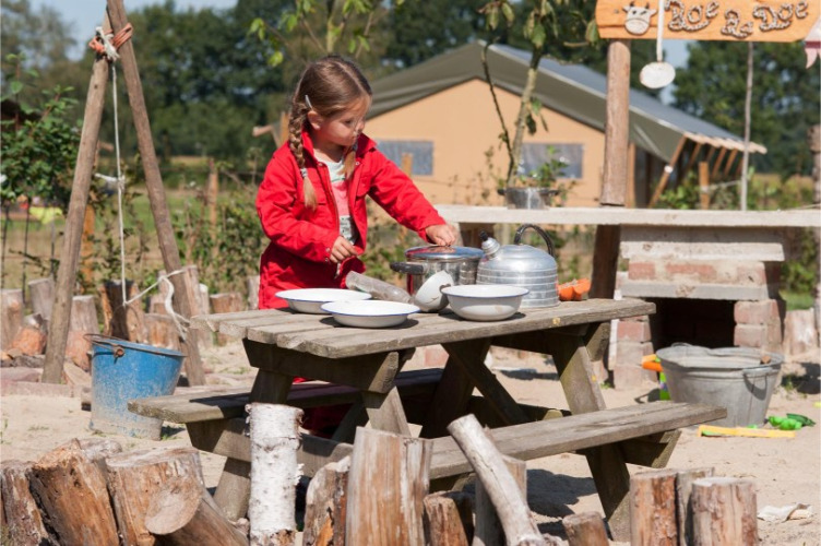 Mädchen im roten Anzug spielt draußen an einem Holztisch im FarmCamps BoeBaDoe in Limburg, Niederlande.