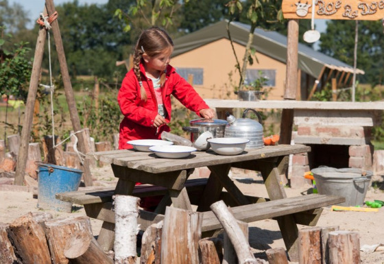 Mädchen im roten Anzug spielt draußen an einem Holztisch im FarmCamps BoeBaDoe in Limburg, Niederlande.