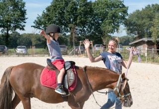 Et barn rider på en pony med hjelm, mens en instruktør smiler og giver high-five på FarmCamps BoeBaDoe.