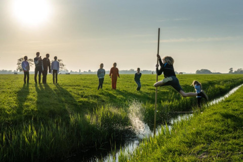 Niños saltan un arroyo con una pértiga en un campo soleado en FarmCamps De Bonte Fries en Friesland, Países Bajos.