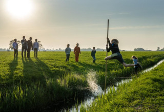 Kinderen springen met een stok over een greppel op het zonnige veld van FarmCamps De Bonte Fries in Friesland.