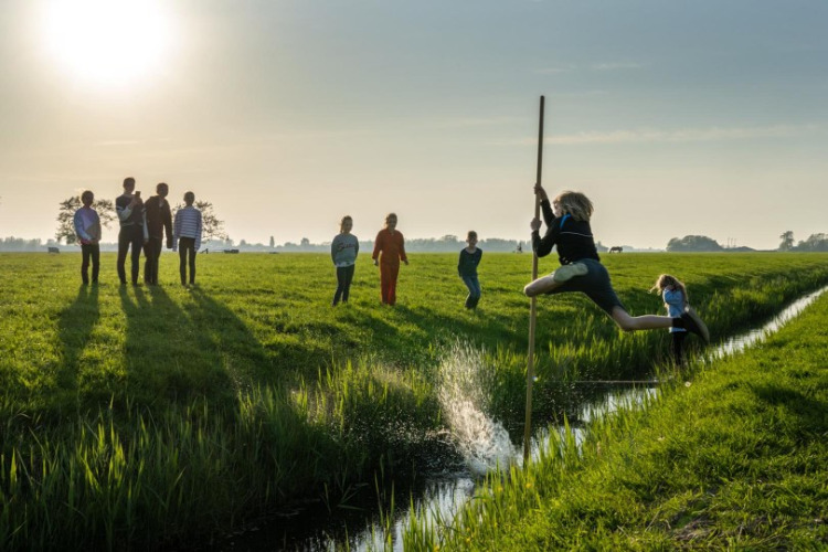 Des enfants sautent un fossé à l’aide d’une perche dans un champ ensoleillé à FarmCamps De Bonte Fries, Frise.
