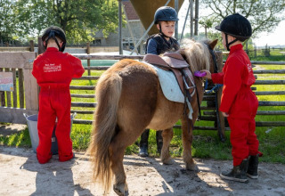 Kinder in roten Overalls und Helmen pflegen ein Pony im Ferienpark FarmCamps De Bonte Fries, Friesland.