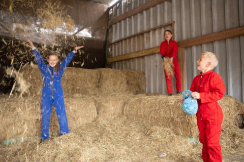 Niños en monos de colores juegan con paja en un granero en FarmCamps De Bosrand, parque vacacional en Drenthe.