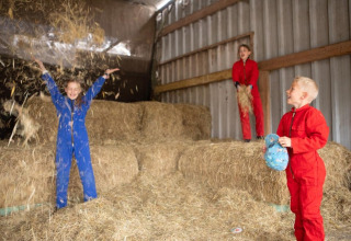 Kinderen in gekleurde overalls spelen met strobalen in een schuur bij FarmCamps De Bosrand in Drenthe.