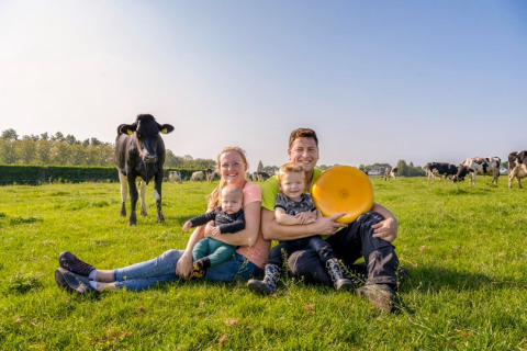 En familie sidder på græsset ved FarmCamps De Buitenhoeve i Gelderland med køer og ost i baggrunden.