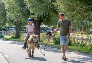 Een man wandelt met een pony waarop een kind zit op een boomrijk pad in een vakantiepark.