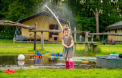 Et barn leger glad i en vandpyt med legetøj og vandstråler på FarmCamps De Geele Bosch i Friesland, Holland.