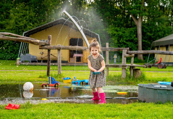 Et barn leger glad i en vandpyt med legetøj og vandstråler på FarmCamps De Geele Bosch i Friesland, Holland.
