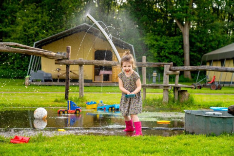 A happy child plays with toys and water on a grassy field at FarmCamps De Geele Bosch in Friesland, Netherlands.