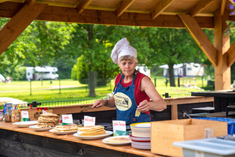 Kvinde i kokkehue serverer pandekager udendørs på FarmCamps De Geele Bosch i Friesland, Holland.