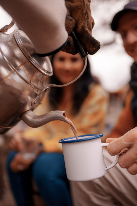Una persona sirve una bebida caliente de una tetera en una taza esmaltada, dos personas al fondo difuminadas.