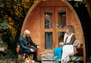Zwei Personen entspannen vor einer Holzhütte im Wald im Ferienpark RØSTIG Dorst in Nordbrabant, Niederlande.
