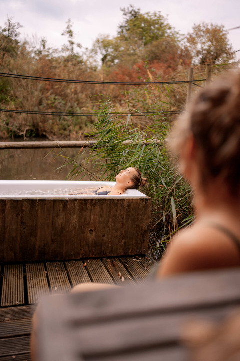 Due donne si rilassano accanto a una vasca da bagno all'aperto immersa nella natura al RØSTIG Dorst, Paesi Bassi.