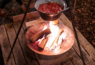 Outdoor-Kochen am Lagerfeuer mit Eintopf im Topf, Brot und Käse auf Holztisch, zwei Frauen im Hintergrund.