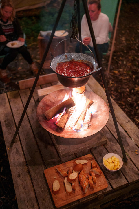 Outdoor cooking at campfire with stew in a pot, bread and cheese on a rustic table, two women in background.