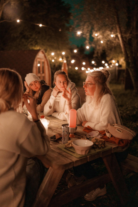 Cuatro mujeres conversan en una mesa de madera iluminada en el parque vacacional RØSTIG Dorst en North-Brabant.
