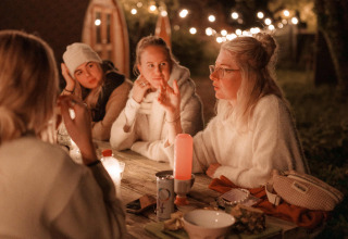 Quattro donne conversano di sera a un tavolo di legno con luci al parco vacanze RØSTIG Dorst, North-Brabant.