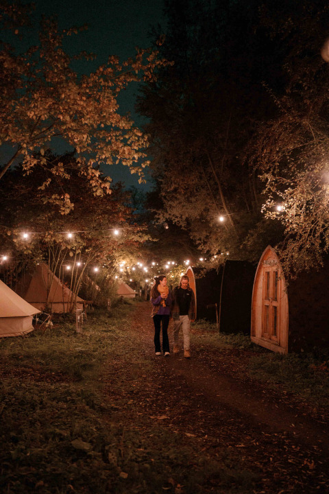 Dos personas caminan bajo guirnaldas de luces entre tiendas glamping por la noche en RØSTIG Dorst, Brabante Septentrional.