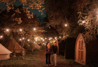 Twee mensen wandelen ‘s avonds onder sfeerverlichting naast glampingtenten in RØSTIG Dorst, Noord-Brabant.