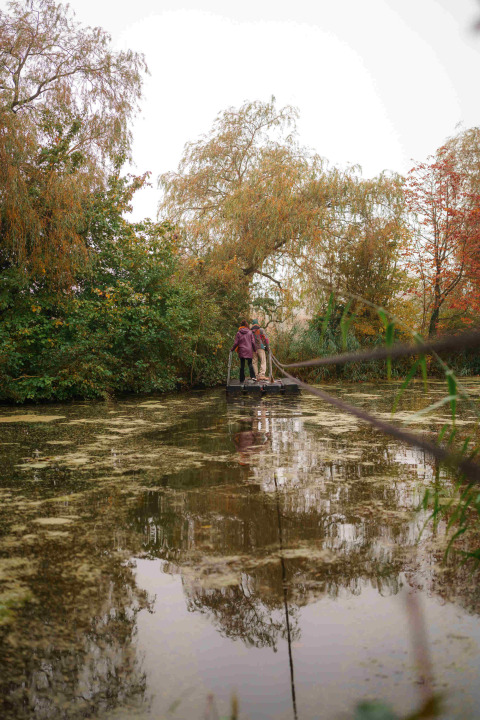 Two people standing on a wooden raft by a pond, surrounded by autumn trees at RØSTIG Dorst, Netherlands.