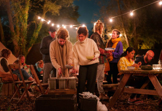 Avondbijeenkomst in RØSTIG Dorst vakantiepark, mensen dineren onder lichtslingers in het bos.