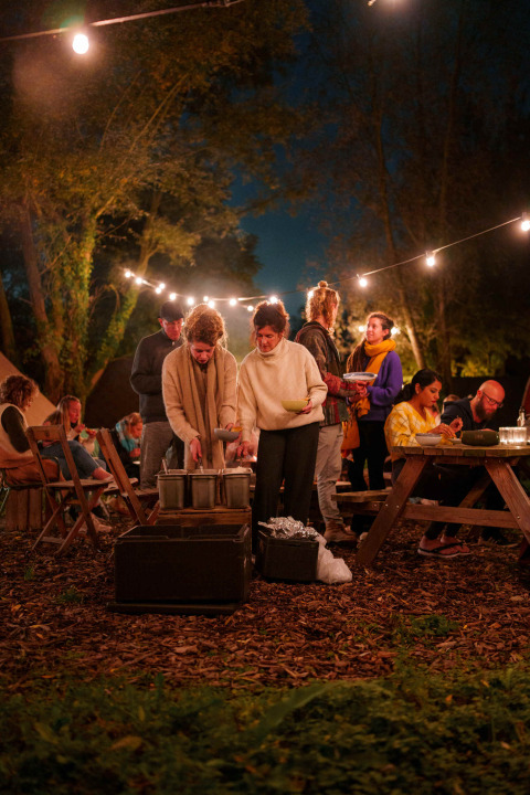 Outdoor evening gathering at RØSTIG Dorst holiday park with people dining under string lights in a forest.
