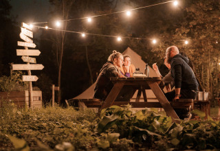 Vier mensen zitten aan een picknicktafel onder sfeerverlichting in RØSTIG Dorst, vakantiepark in Noord-Brabant.