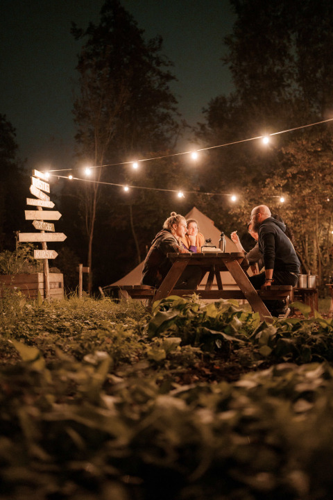 Cuatro personas se sientan en una mesa de picnic bajo luces en RØSTIG Dorst, un parque vacacional en North-Brabant.