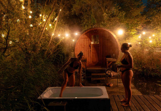 Twee vrouwen in badkledij genieten bij een buitenbad en sauna, sfeervol verlicht in het bos tijdens de nacht.