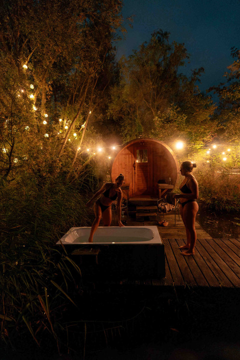 Two women in swimsuits relax by an outdoor bathtub and sauna, surrounded by lights in a forest at night.