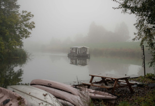 Mistig tafereel bij vakantiepark RØSTIG Dorst, Nederland, met kano’s aan de oever en woonboot op het meer.