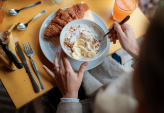 Ontbijt met croissant, yoghurt met banaan en granola aan een gele tafel in RØSTIG Dorst vakantiepark.