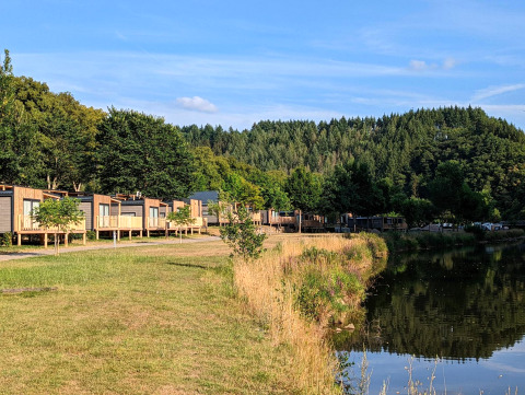 Glamping-accommodatie aan een meer met moderne huisjes, groene bomen en uitzicht op bosrijke heuvels.