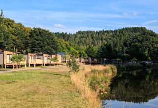 Glamping-accommodatie aan een meer met moderne huisjes, groene bomen en uitzicht op bosrijke heuvels.