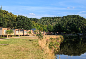 Glamping-Unterkünfte am See mit modernen Hütten, Bäumen und Blick auf einen Wald an einem Sommertag.