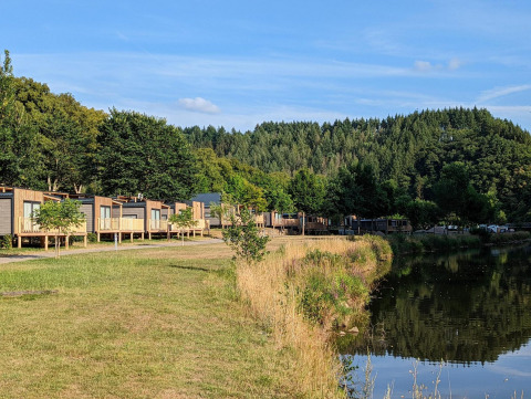 Hébergements glamping au bord d’un lac, cabanes modernes, arbres verts et vue sur une forêt vallonnée.