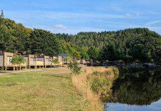 Hébergements glamping au bord d’un lac, cabanes modernes, arbres verts et vue sur une forêt vallonnée.
