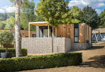 Moderna cabaña Domus con fachada de madera, muro de piedra y terraza, rodeada de verde paisaje natural.