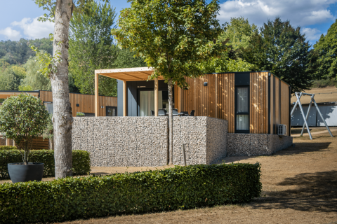 Moderne Domus Lodge mit Holzverkleidung, Steinmauer und Terrasse in grüner, naturnaher Umgebung.
