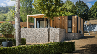 Lodge Domus moderne avec façade en bois, mur en pierre et terrasse au cœur d’un environnement verdoyant.