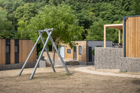 Außenaufnahme der Domus-Lodge mit modernen Holzbauten, Steinzäunen, Schaukel und viel Grün.