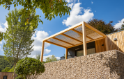 Modern Domus lodge at Kohnenhof in Luxembourg with wooden pergola, stone wall, and lush trees on a sunny day.