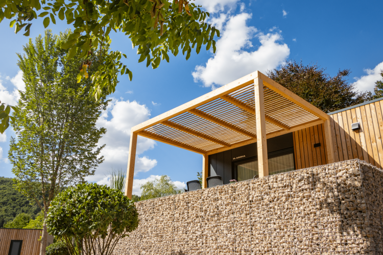 Lodge moderne Domus à Kohnenhof, Luxembourg, avec pergola en bois, mur en pierre et arbres sous ciel bleu.